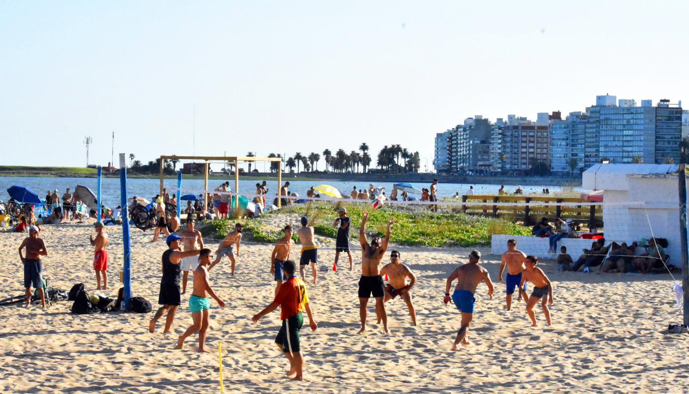 playa en dia soleado gran cantidad de personas realizando actividades deportivas, personas tomando un baño. Al fondo los edificios del Barrios de gran altura 