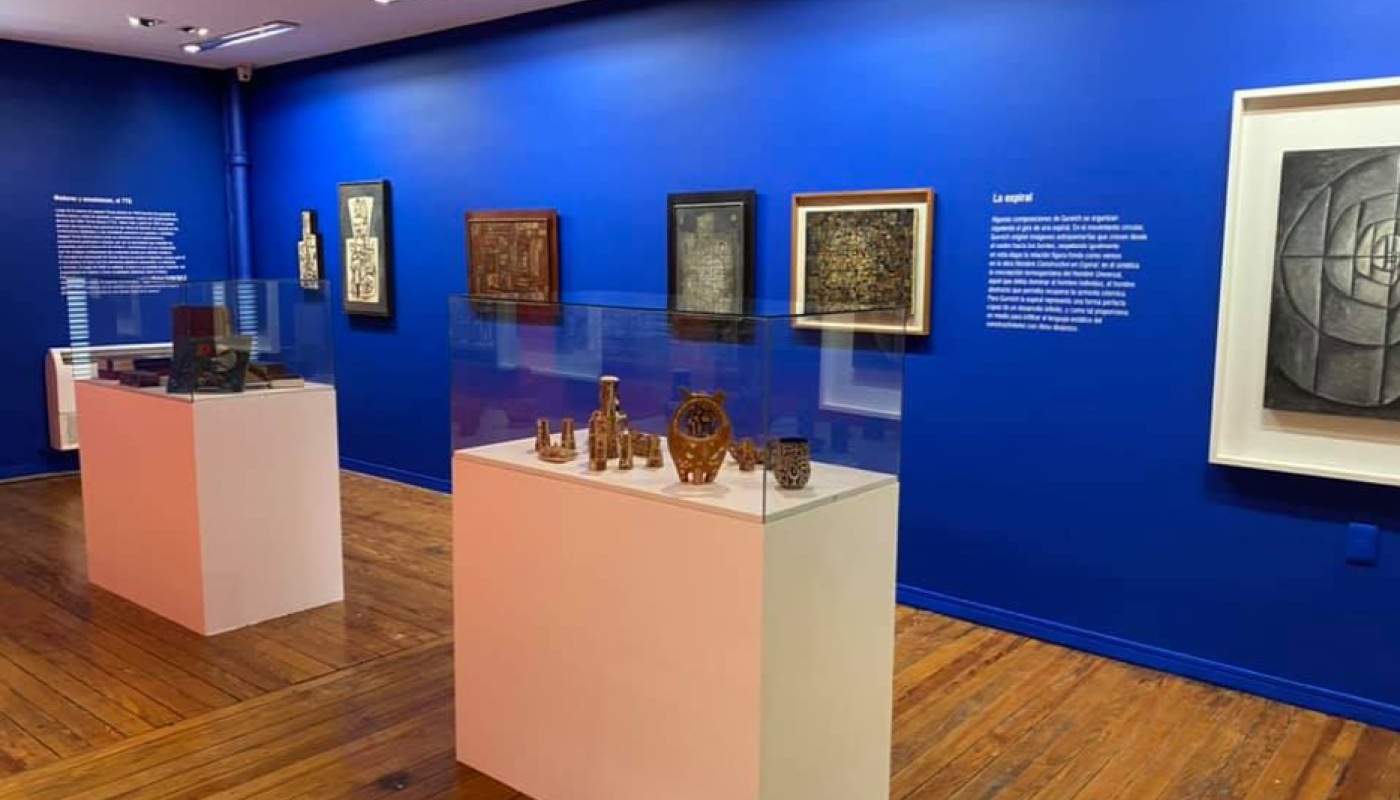 Foto parcial de la sala. Paredes azul fuerte con cuadros colgados. Exhibidores cuadrados, blancos con campanas de cristal y objetos exhibidos. En las paredes hay textos explicativo de las obras.