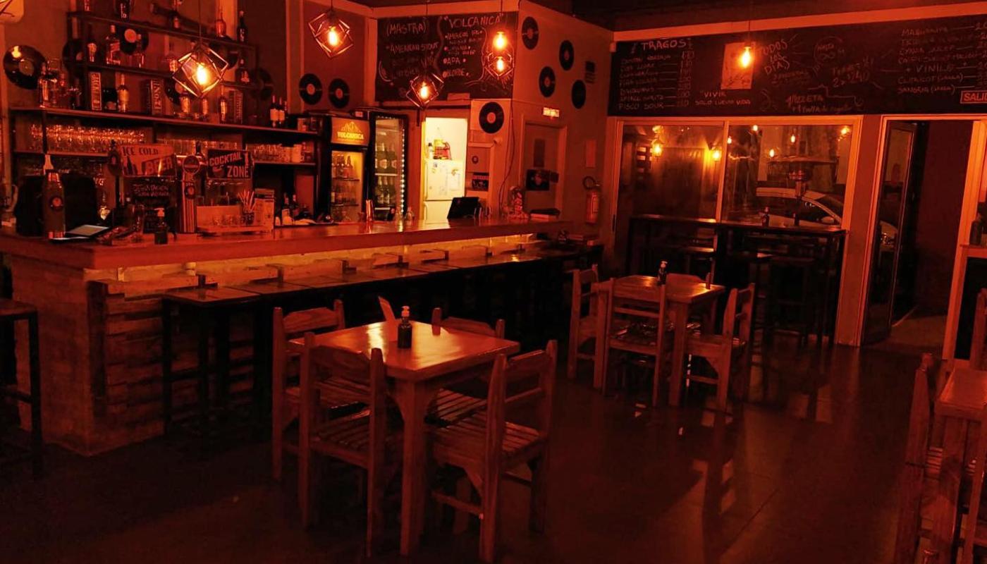 Foto nocturna del salón del local con mesas de madera y sillas y la barra al fondo con discos de vinilo como decoración  