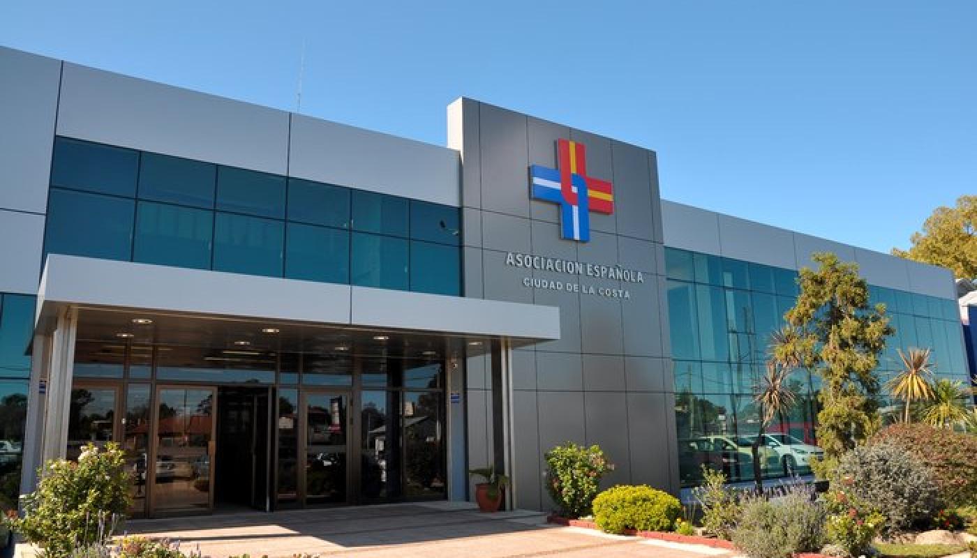 Una de las sedes del hospital en un día soleado. Construcción moderna de color gris con mucho vidrio templado. Logo de la empresa en la fachada en color azul y rojo. Pasto y arboles al frente.
