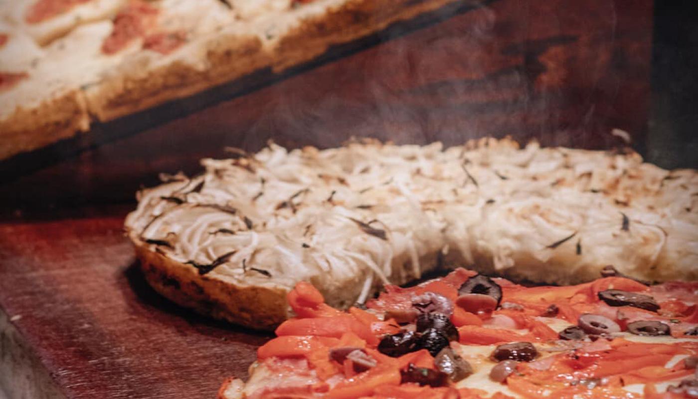 Foto con una pizza con tomate y aceitunas y una fugazzeta, en el fondo se observan más trozos de pizza 