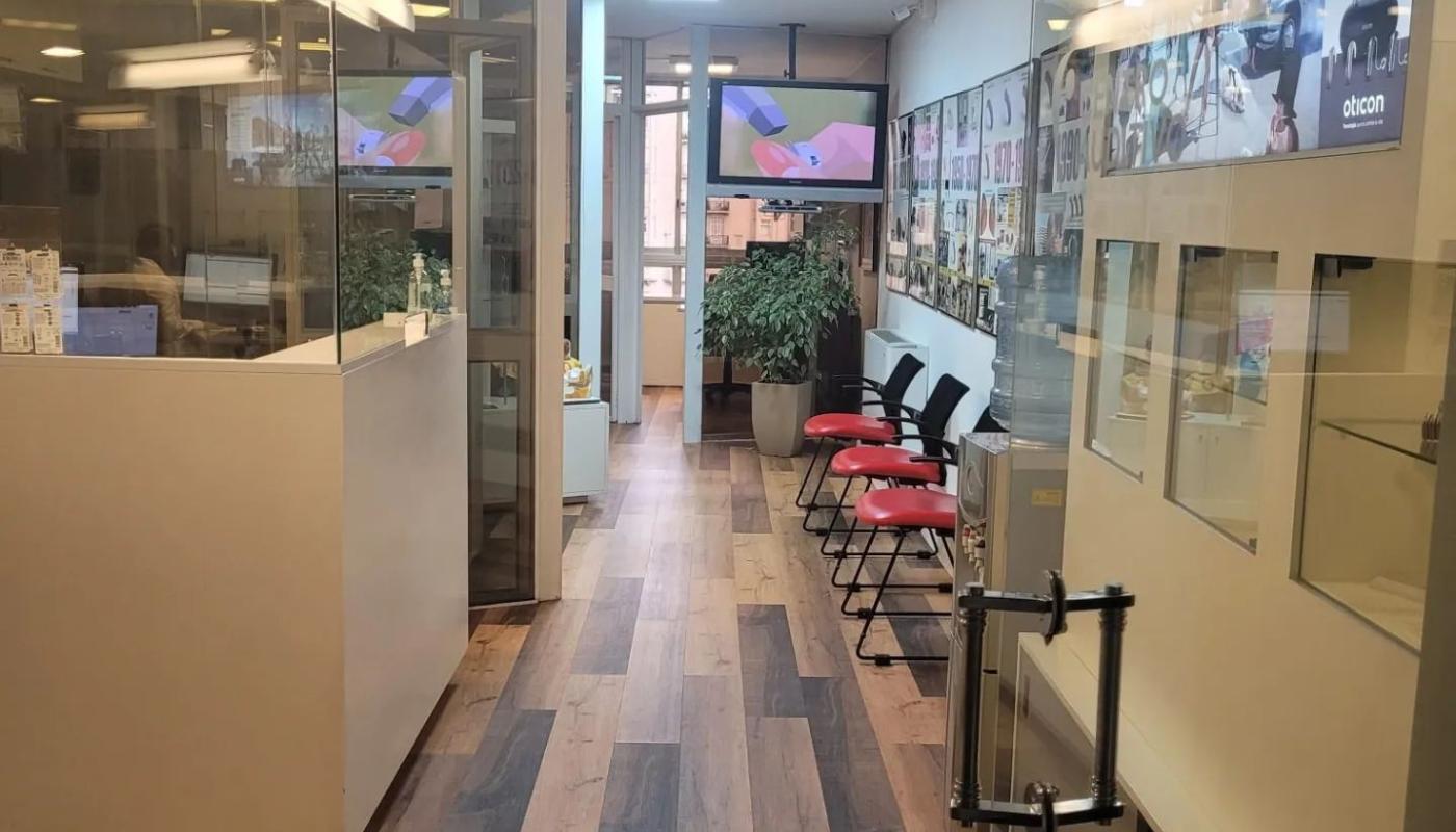 Sala de espera con piso de madera y escritorio de recepción. Mucho vidrio. Cartelería alusiva. 