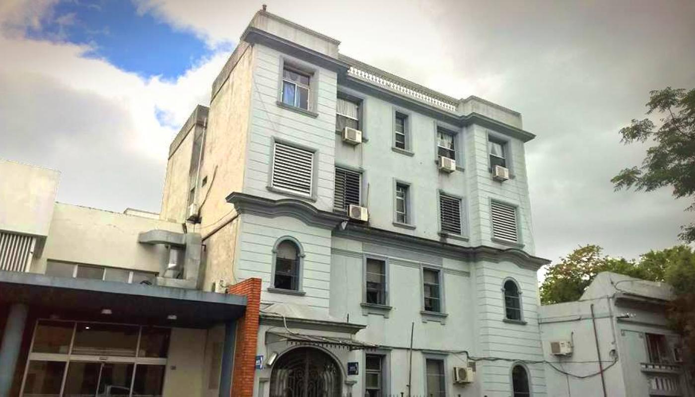 Edificio antiguo de color blanco de dos pisos. Entrada vidriada en forma de arco. 