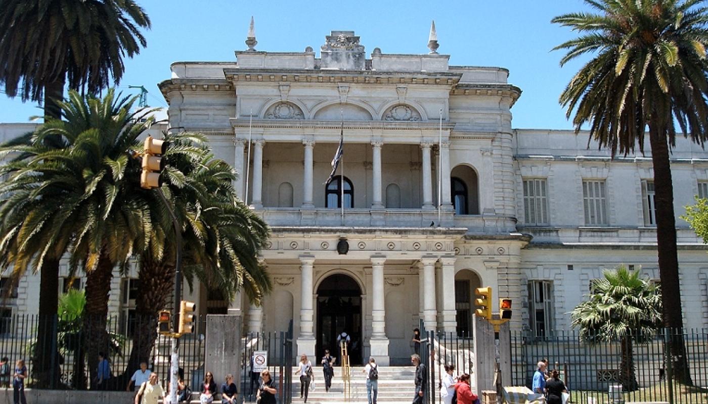 Día soleado con el edificio al fondo y dos palmeras al frente. Casona antigua de dos pisos blanca sobre la Av 8 de Octubre. Ingreso con escalinata pequeña y fachada con columnas. Segundo piso con balcón y columnas. 