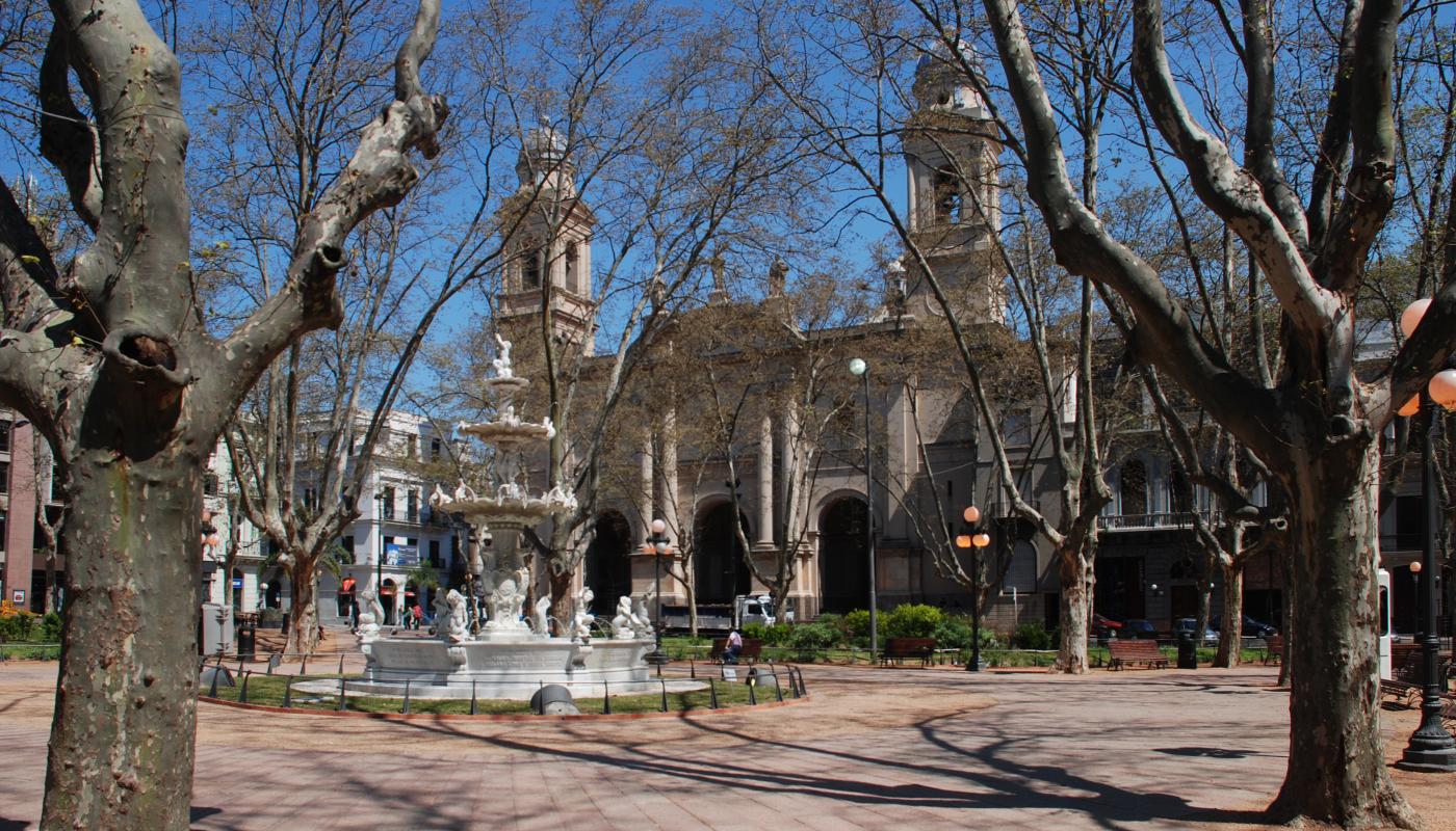 Debajo de unos árboles que han perdido sus hojas, hay una fuente blanca, antigua, de varios niveles. Al fondo se ve la Iglesia Matriz.