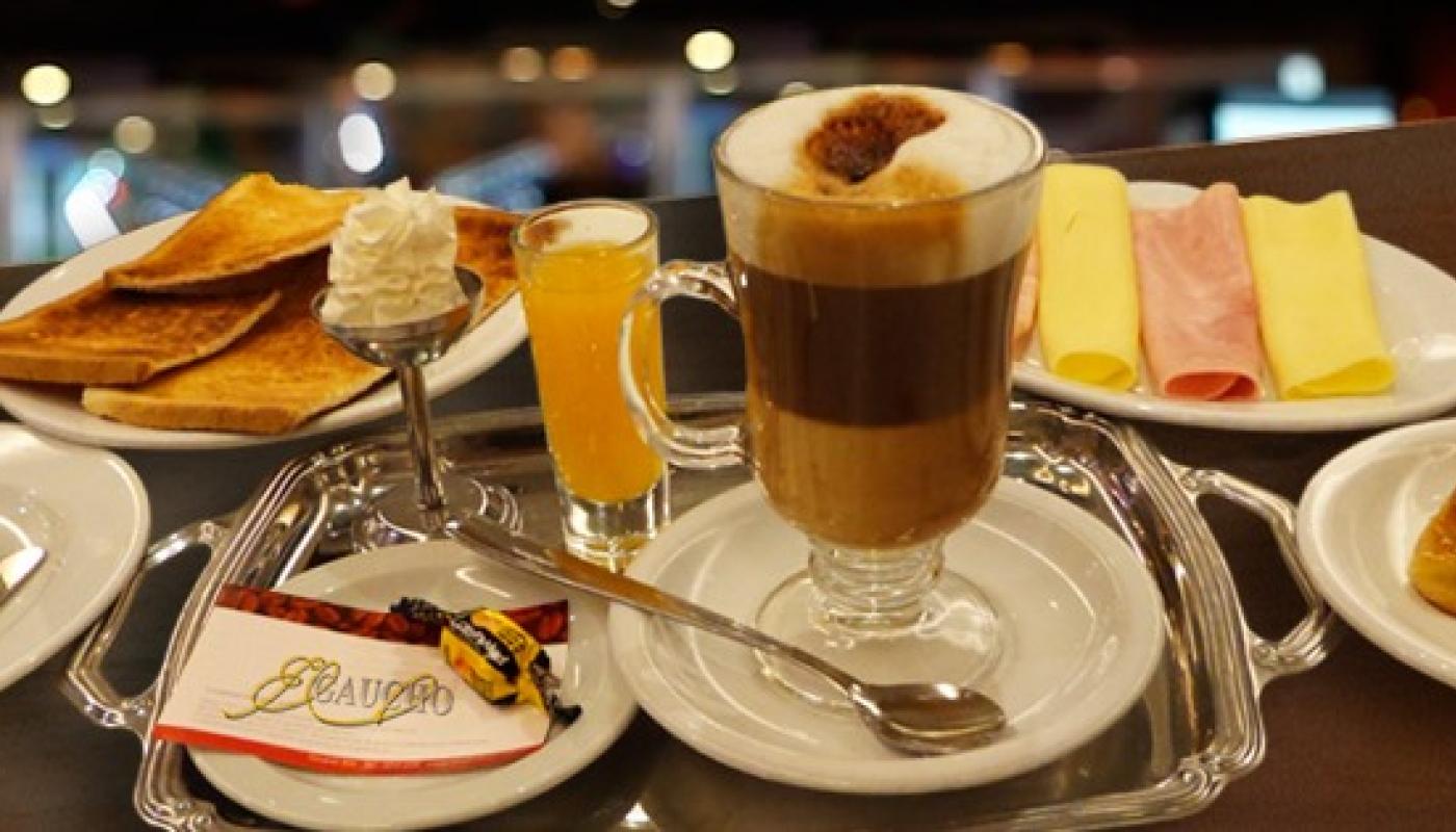 Mesa completa con la merienda que tiene capuchino, medialunas, tostadas con mermelada y queso de untar y fiambres,