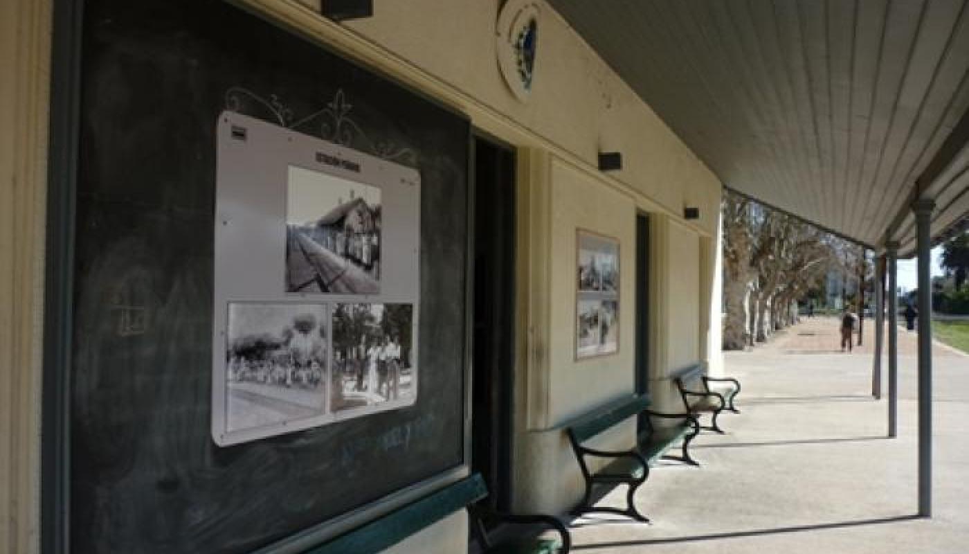 Andén de una estación de tren. Hay dos bancos antiguos de hierro y madera, debajo de un alero. Al fondo hay una fila de árboles.