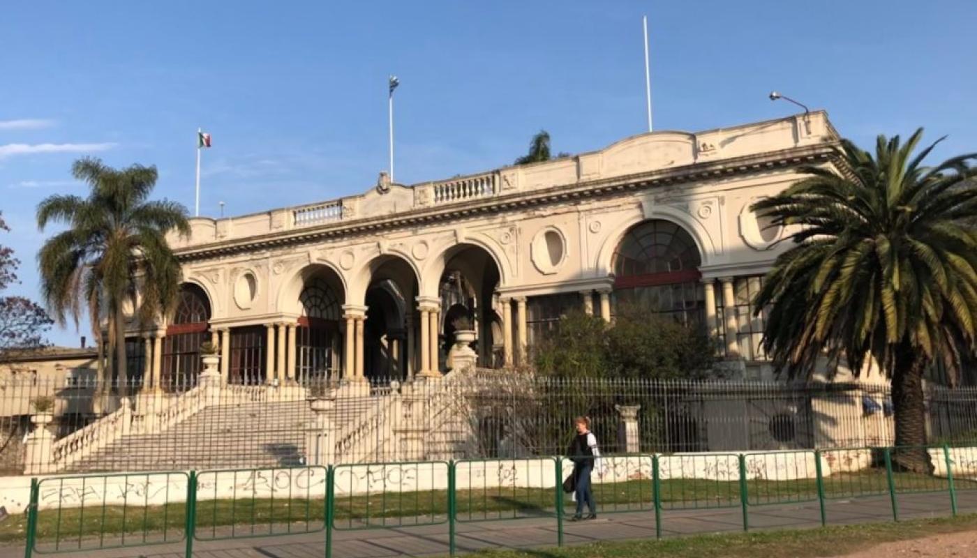 Foto completa del edificio del hospital con reja alrededor y jardín al frente. Edificio antiguo de estilo neoclásico con escalinata central en mármol. Hall de recepción con columnatas y arcos. Techo a la porteña con un único piso. Falsos ojo de buey en la fachada. 