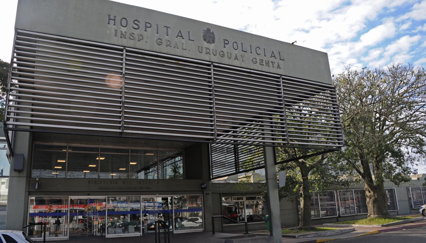 Edificio moderno gris metalizado. Fachada en forma de cortina metálica con el nombre impreso del hospital "Hospital Policial". Ingreso netamente vidriado. 