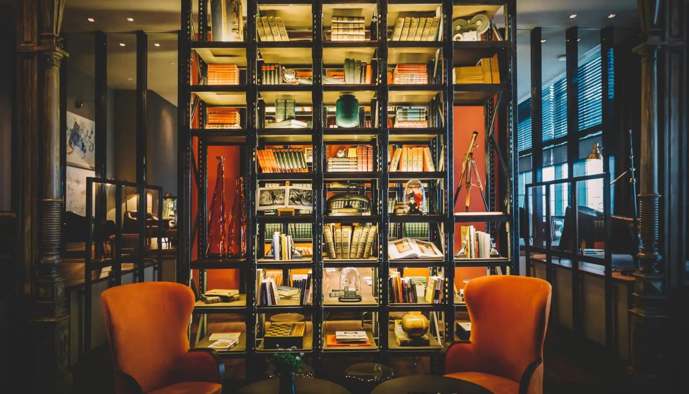  Interior del Hotel Hyatt Centric. Gran estructura negra con estantería abierta. Hay libros antiguos, cámara de video antigua, jarrones, caja de madera de juego de ajedrez, dos telescopios. Frente a la estantería hay dos sillones naranjas antiguos y dos mesitas negras redondas muy bajas.