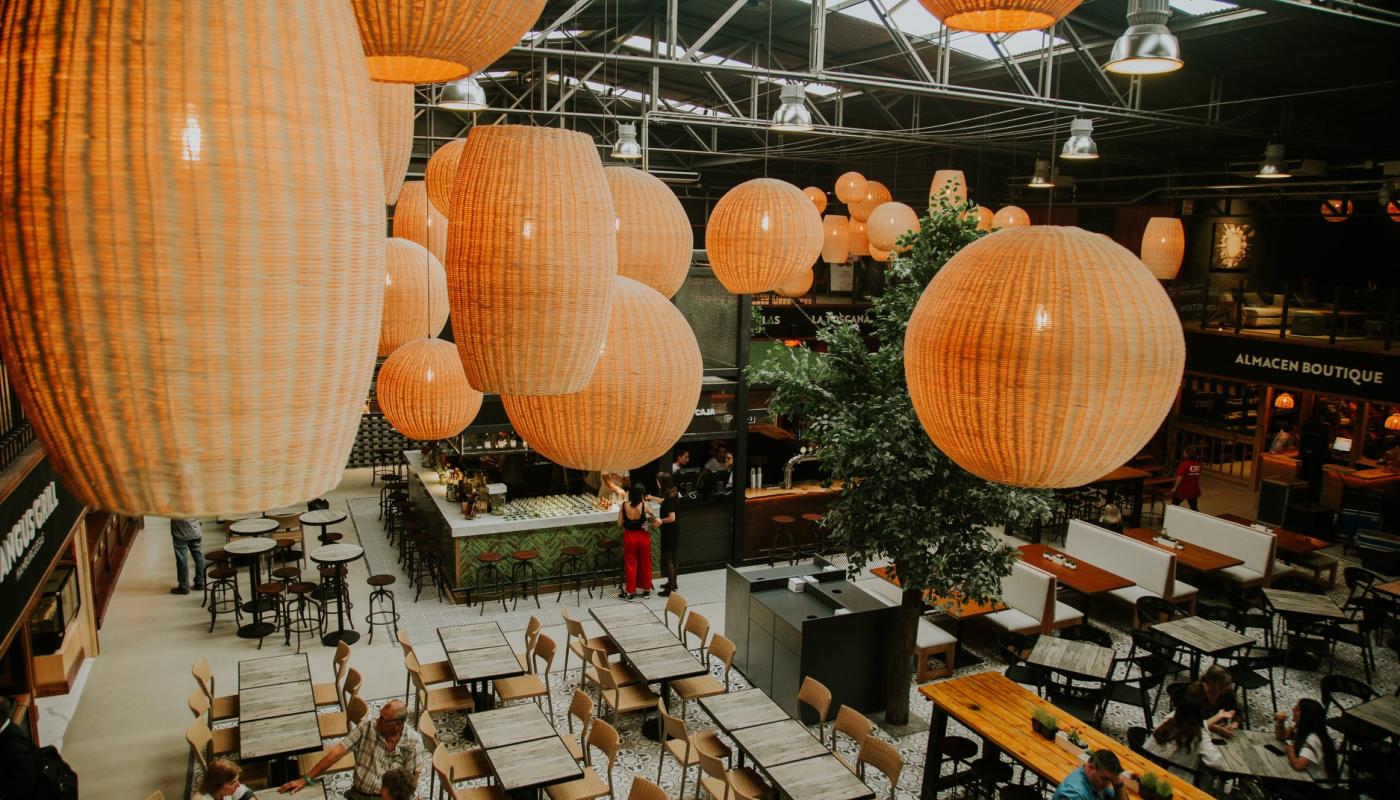 Foto desde arriba de interior del mercado destancando sus lamparas tipo mimbre ovales y redondas, gran plaza de comidas con mesas del mismo color en madera clara y barras de locales gastronomicos alrededor