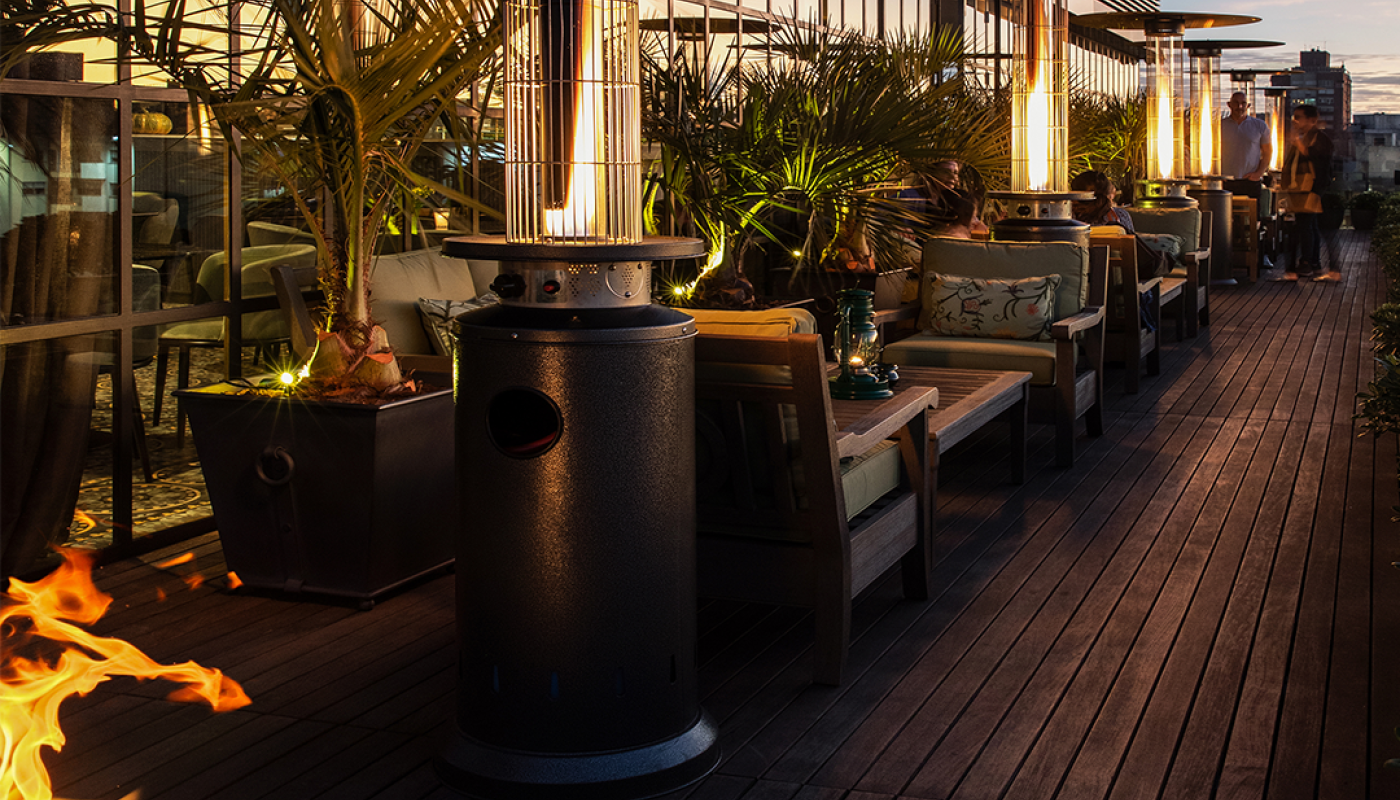 Área exterior del Hotel Montevideo sobre un deck de madera. Zona de sillones encontrados y mesas bajas. Entre ellos, calefactores de exterior con tubos verticales encendidos para calefaccionar. Detrás, la fachada vidriada.