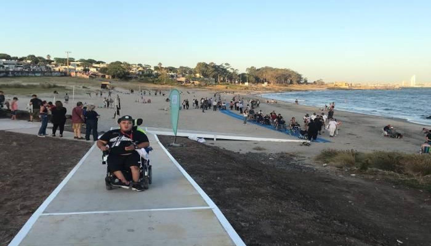 playa accesible con rampa hacia la arena persona en silla de ruedas ene lla, al fondo la arena y el mar