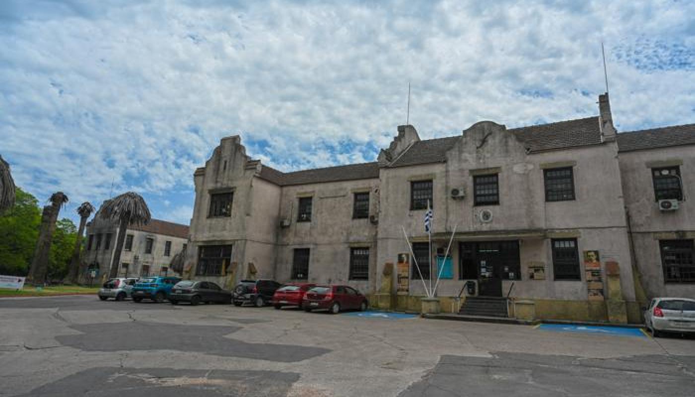 Edificio antiguo de dos pisos de color claro. Localizado en un parque. Detalles arquitectónicos en techos y alerones. 