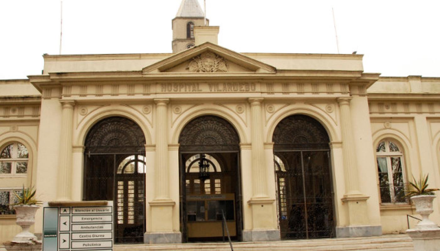 Edificio principal del Hospital en estilo neoclásico de color claro. Fachada principal con 3 arcos y lintel en triangulo. El nombre está escrito en la fachada en letras mayúsculas. Detrás se ve una torre alta.  