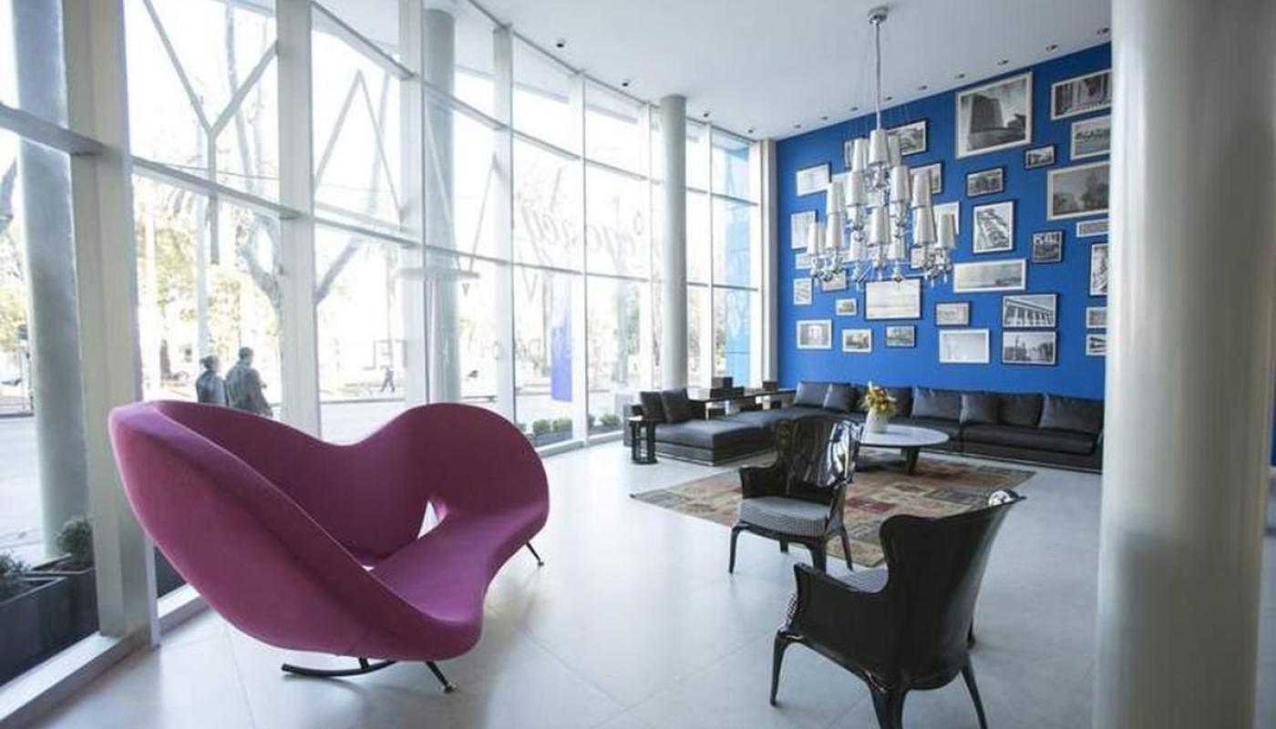 lobby del Hotel. Hay sillones de diversos tamaños y colores. De costado hay una pared todo vidriada y al fonfo una pared de color azul intenso con muchos cuadros en blanco y negro de fotos antiguas