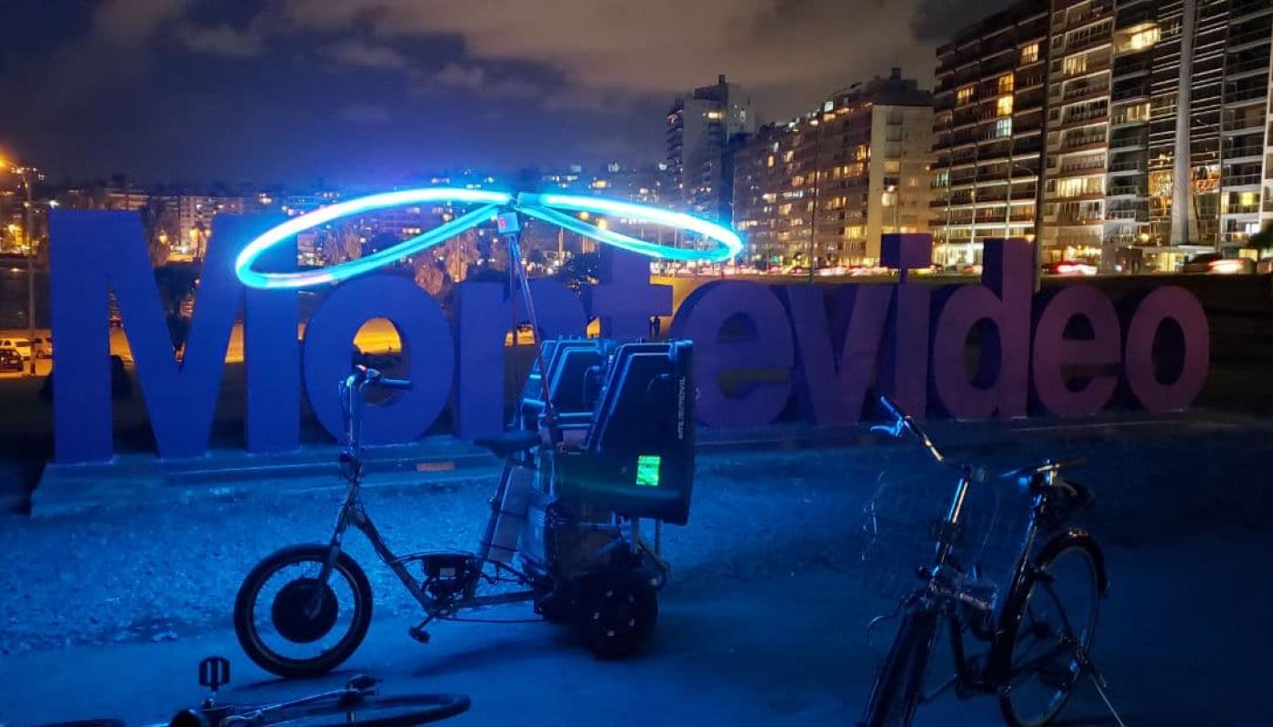 Foto nocturna del Cartel de Montevideo con una bicicleta iluminada con un aro de luz y parlantes