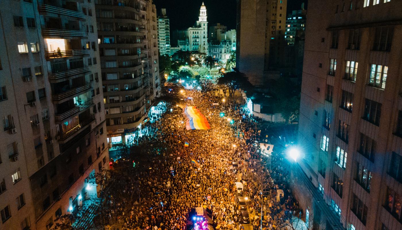 "Vista aérea nocturna de la Marcha de la Diversidad en Montevideo, con una multitud llenando la Avenida 18 de Julio y una gran bandera arcoíris extendida en el centro, iluminada por las luces de la ciudad y con el Palacio Salvo visible al fondo.