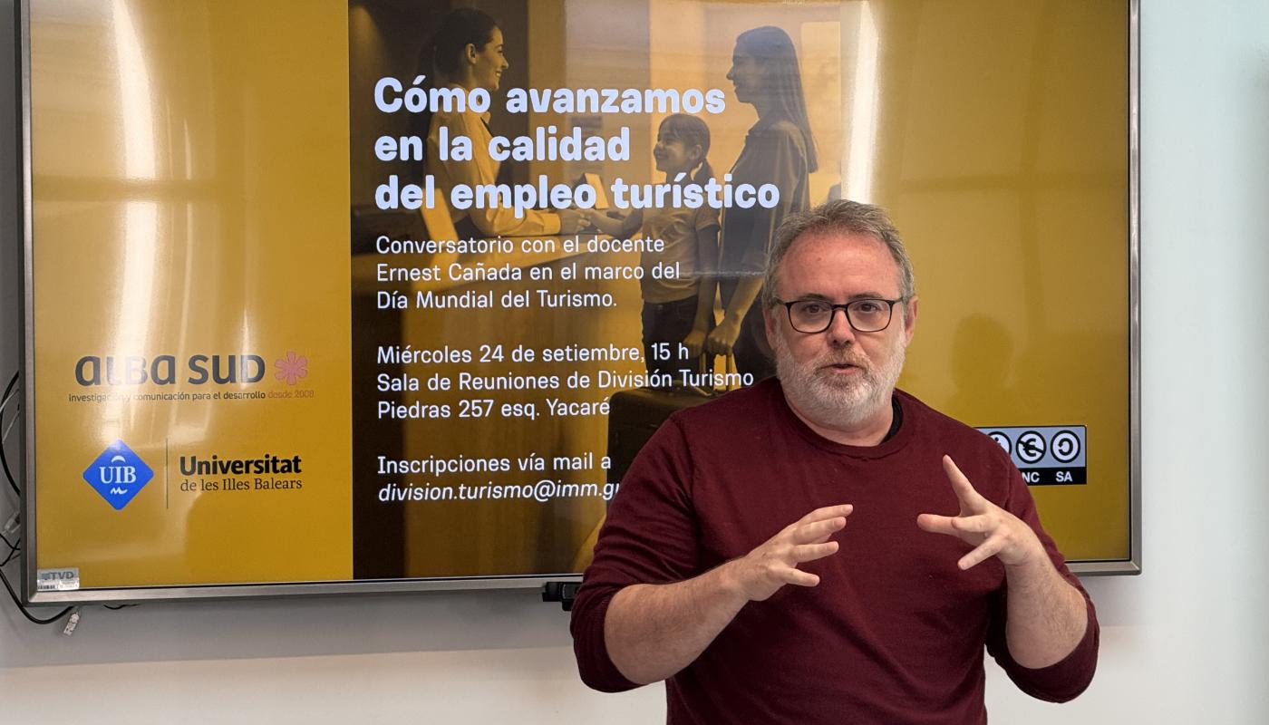 Docente en primer plano gesticulando mientras explica, al lado de una pantalla donde se lee la convocatoria al conversatorio sobre empleo turístico, con logos de instituciones académicas y de turismo.