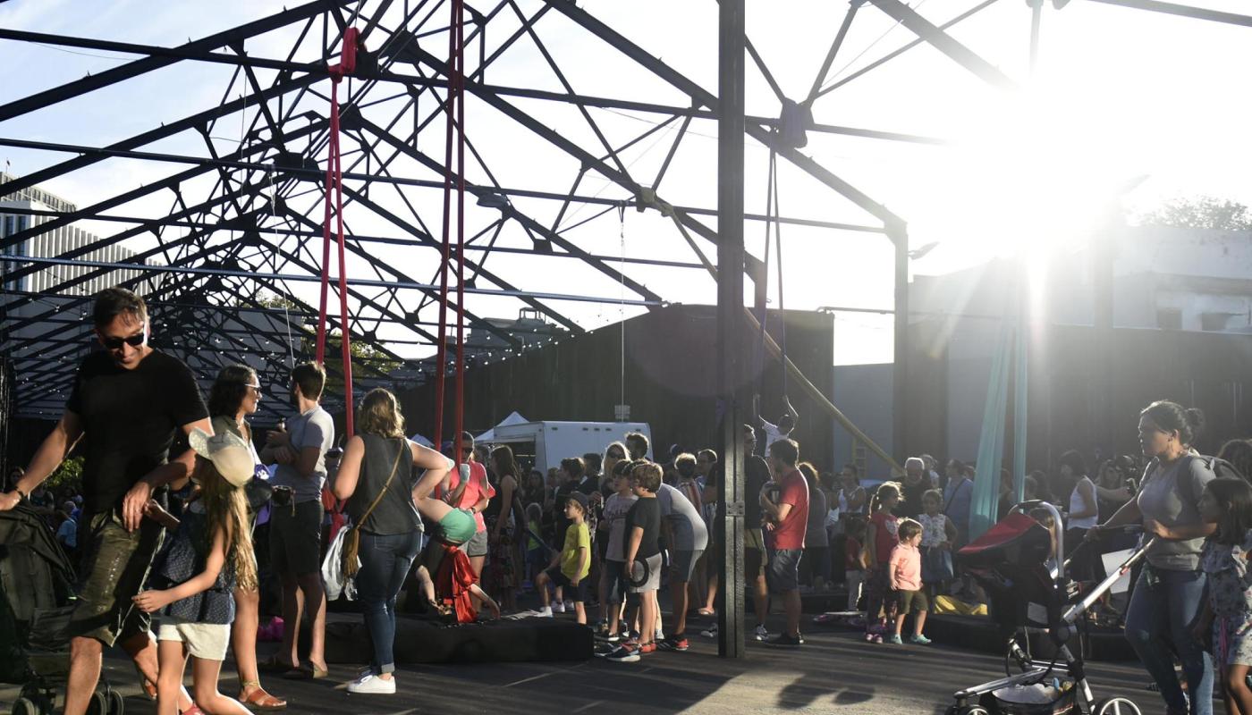 Espacio abierto con estructura metálica de techo sin cubrir, lleno de familias y niños disfrutando de actividades recreativas al atardecer. Algunas personas caminan, otras empujan cochecitos y varios niños juegan cerca de columpios y telas acrobáticas, con la luz del sol entrando desde el fondo.