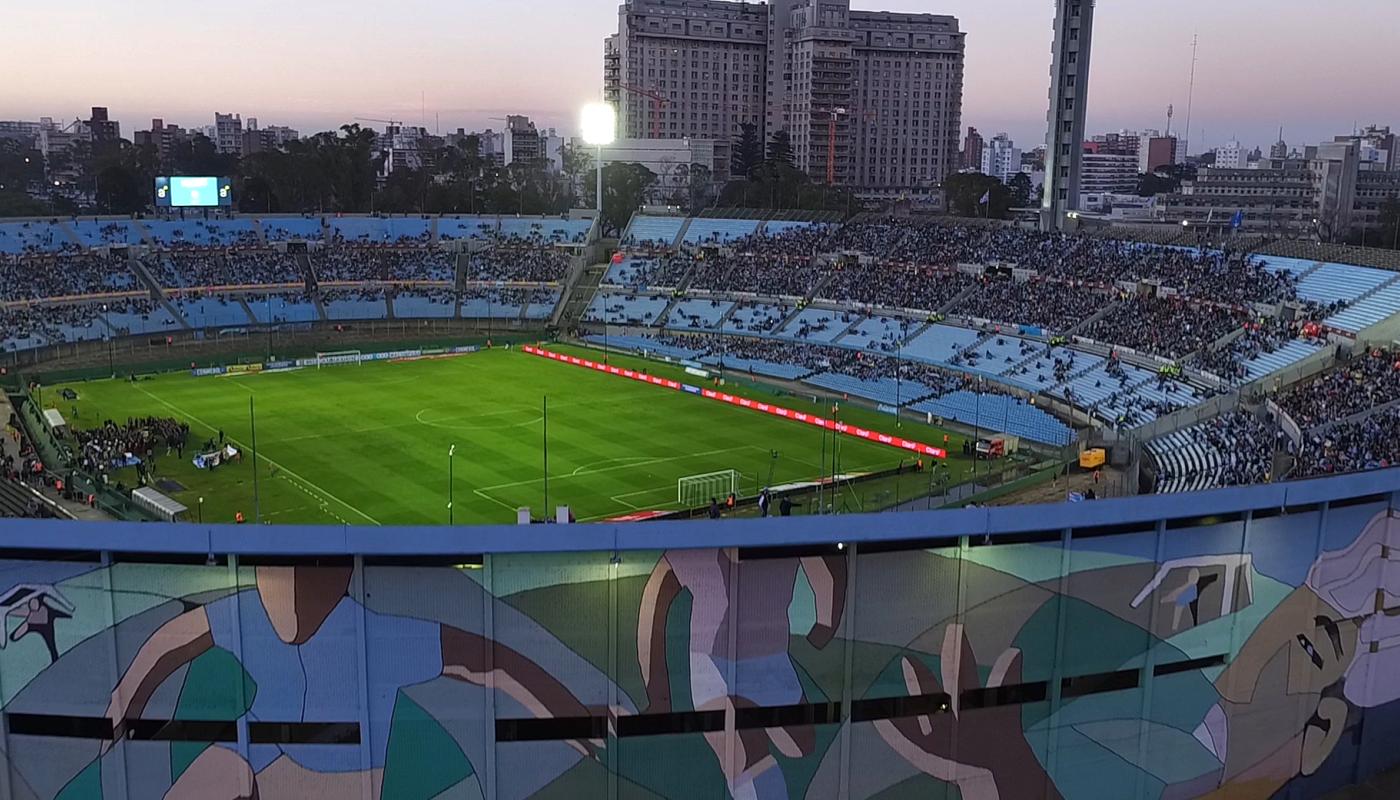 Vista panorámica del Estadio Centenario en Montevideo al atardecer, con las tribunas parcialmente colmadas de público, el campo de juego iluminado y un mural deportivo en primer plano.