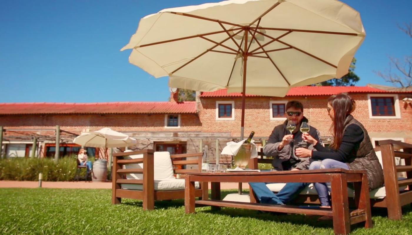 Pareja sentada al aire libre en sillones de madera con cojines, bajo un gran paraguas blanco. Brindan con copas de vino frente a la fachada de ladrillo de la Bodega Valdi, en un día soleado.