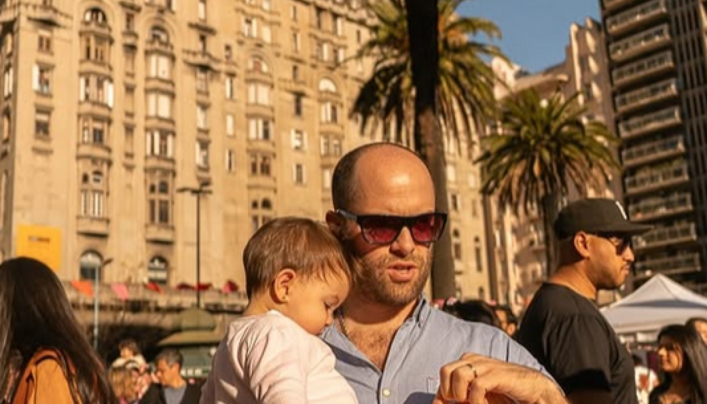 Hombre con gafas de sol sostiene en brazos a una niña pequeña mientras come en la Plaza Independencia de Montevideo. De fondo se ven el Palacio Salvo, palmeras y varias personas disfrutando de una jornada al aire libre.