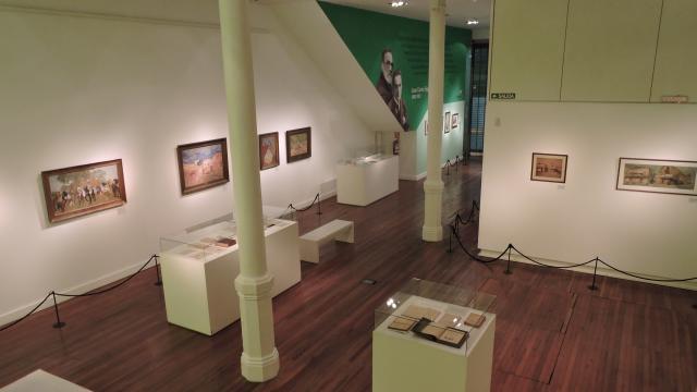 Foto parcial de una de las salas. Paredes pintadas de verde clarito. Columnas en el medio finitas y pequeñas. Cuadros exhibidos en las paredes. Piso de madera. Exhibidores cuadrados, blancos y con campana de vidrio en el centro de la sala, hay 3 exhibidores. Y un banco blanco para sentarse
