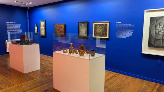 Foto parcial de la sala. Paredes azul fuerte con cuadros colgados. Exhibidores cuadrados, blancos con campanas de cristal y objetos exhibidos. En las paredes hay textos explicativo de las obras. 