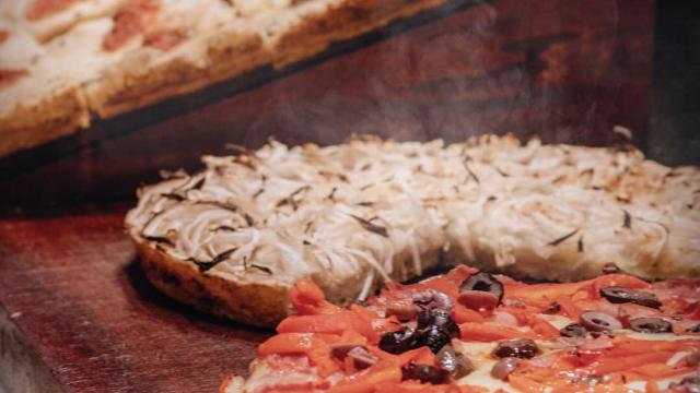 Foto con una pizza con tomate y aceitunas y una fugazzeta, en el fondo se observan más trozos de pizza 