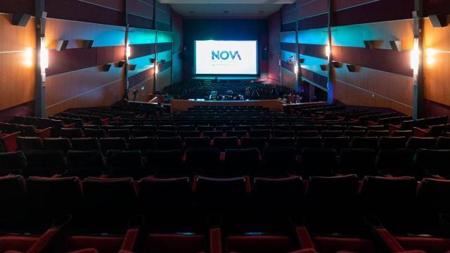 Foto de atrás de la sala con butacas rojas. Escenario al frente con pantalla visible.