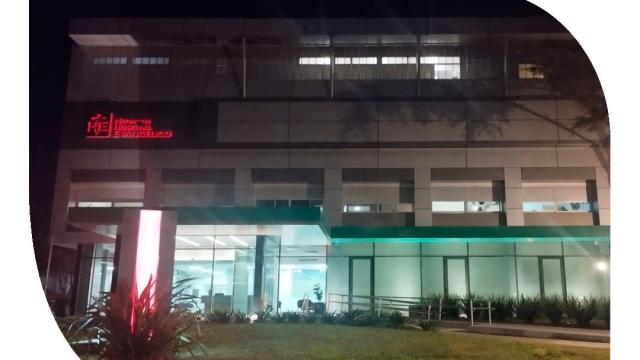 Foto nocturna de la mutualista y su sede principal. Edificio moderno vidriado con jardín al frente. 