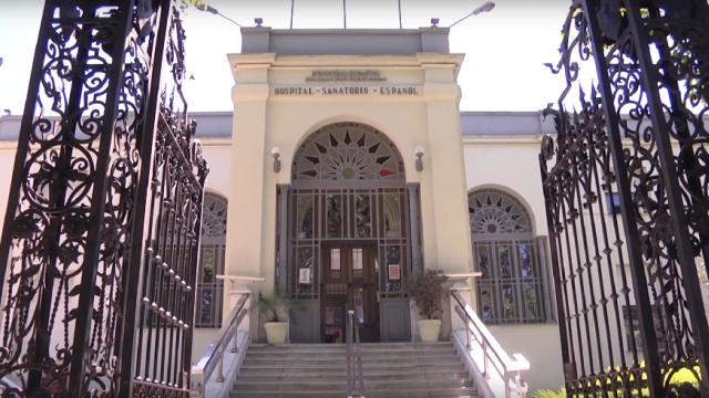 Construcción antigua. Portón de hierro con detalles trabajados en él. Entrada del hospital con escalinata. Cuerpo principal en forma de torre de color beige y vitrales de colores. 