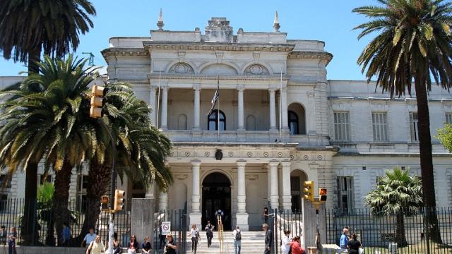 Día soleado con el edificio al fondo y dos palmeras al frente. Casona antigua de dos pisos blanca sobre la Av 8 de Octubre. Ingreso con escalinata pequeña y fachada con columnas. Segundo piso con balcón y columnas. 