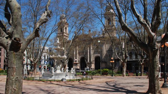 Debajo de unos árboles que han perdido sus hojas, hay una fuente blanca, antigua, de varios niveles. Al fondo se ve la Iglesia Matriz.