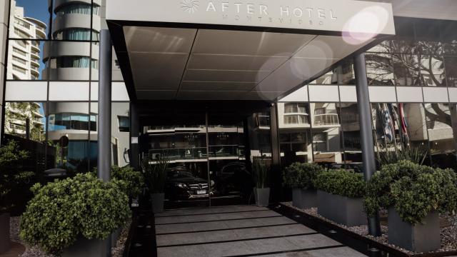 Entrada moderna al hotel. Hay una puerta de vidrio corrediza. Hay plantas y arbustos en el ingreso. 