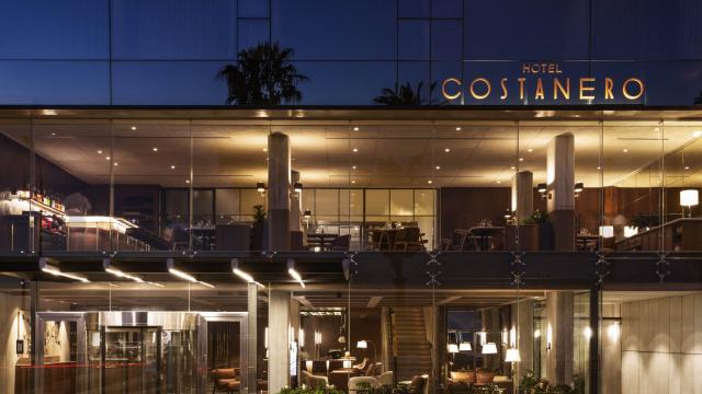 La fachada de cristal del Hotel Costanero deja ver todo el interior en sus dos plantas. En la planta baja hay varias lámparas de pie, sillones individuales, la recepción, la escalera y el ascensor. En la planta superior hay muchas luces dicroicas.   
