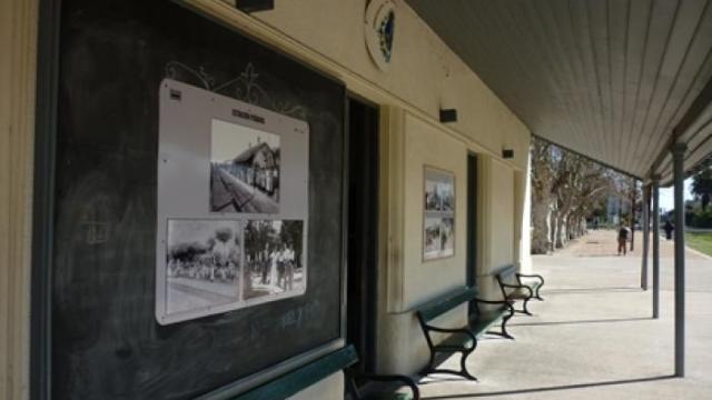 Andén de una estación de tren. Hay dos bancos antiguos de hierro y madera, debajo de un alero. Al fondo hay una fila de árboles.
