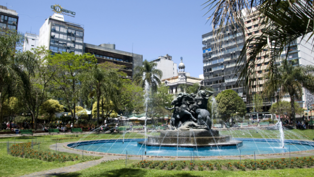 En el centro de una plaza muy verde (con césped y diversos árboles), hay una escultura que representa a algunas personas batallando encima de sus caballos.