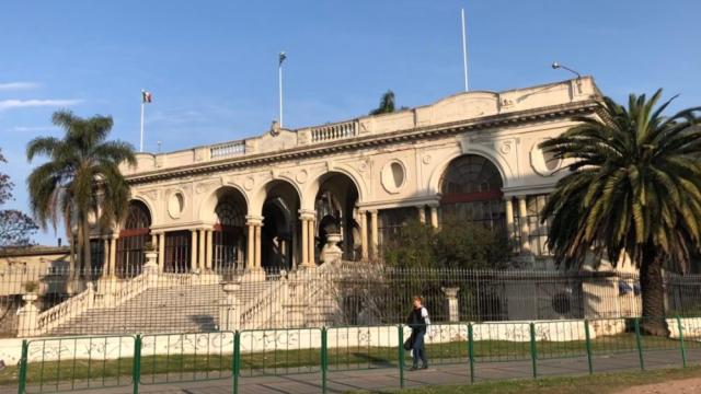 Foto completa del edificio del hospital con reja alrededor y jardín al frente. Edificio antiguo de estilo neoclásico con escalinata central en mármol. Hall de recepción con columnatas y arcos. Techo a la porteña con un único piso. Falsos ojo de buey en la fachada. 
