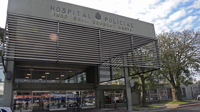 Edificio moderno gris metalizado. Fachada en forma de cortina metálica con el nombre impreso del hospital "Hospital Policial". Ingreso netamente vidriado. 