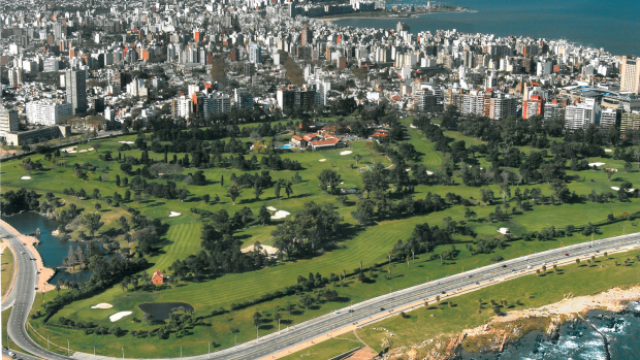 - Vista aérea del Club de Golf de Punta Carretas. Un gran campo verde con mucha vegetación y un lago. Detrás, parte de la ciudad de Montevideo.
