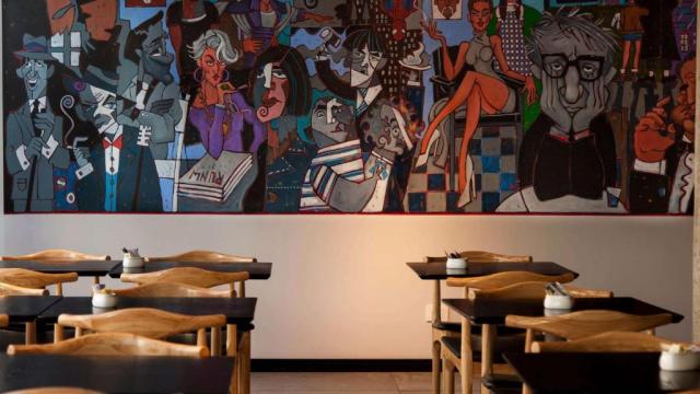 Parte del restaurante del hotel. Hay mesas con sillas de madera. Al fondo, un mural pintado sobre la pared con colores muy vivos, en el que aparecen varias personas en distintas situaciones.