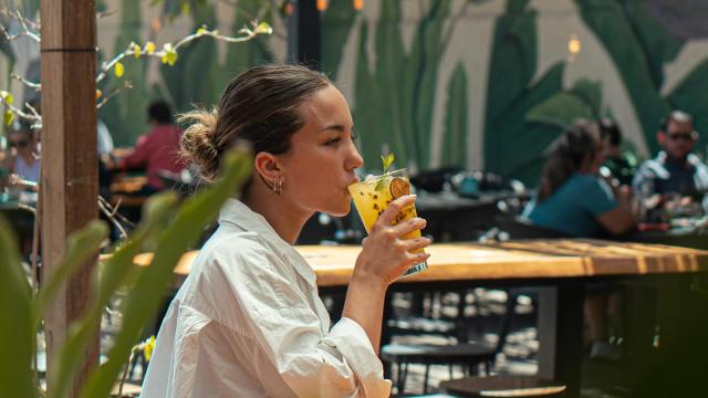 Foto del Jardin del mercado con mesas y sillas, muchas plantas verdes, murales pintados con hojas verdes, chica tomando un trago color amarillo frutal