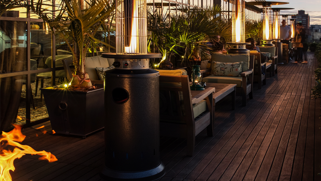 Área exterior del Hotel Montevideo sobre un deck de madera. Zona de sillones encontrados y mesas bajas. Entre ellos, calefactores de exterior con tubos verticales encendidos para calefaccionar. Detrás, la fachada vidriada.