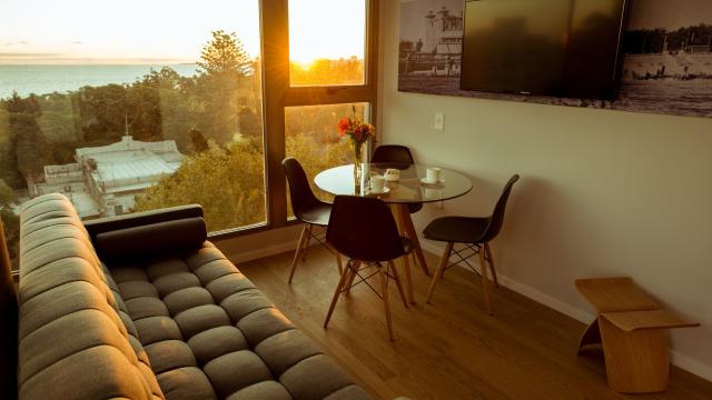 Interior de una habitación. Hay un sillón cama y una mesa redonda de vidrio con cuatro sillas. En la pared cuelga una TV.
