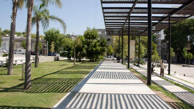 Vista a pie de calle del Parque Seregni. Parte del piso es de césped y parte de cemento. Hay una pared de piedras, diversos árboles y una palmera. También hay una pérgola que hace sombra en el suelo.