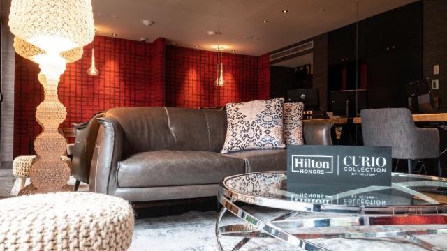 Parte del lobby del hotel. Hay un sillón de dos cuerpos de cuero, una banqueta y una mesa ratona redonda de vidrio. Al fondo hay una barra y sillones altos.   