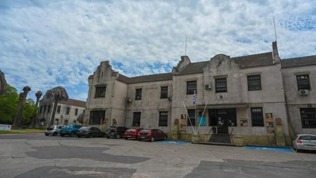 Edificio antiguo de dos pisos de color claro. Localizado en un parque. Detalles arquitectónicos en techos y alerones. 
