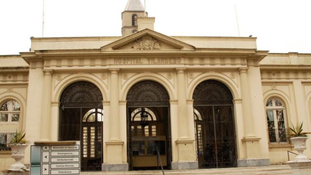 Edificio principal del Hospital en estilo neoclásico de color claro. Fachada principal con 3 arcos y lintel en triangulo. El nombre está escrito en la fachada en letras mayúsculas. Detrás se ve una torre alta.  